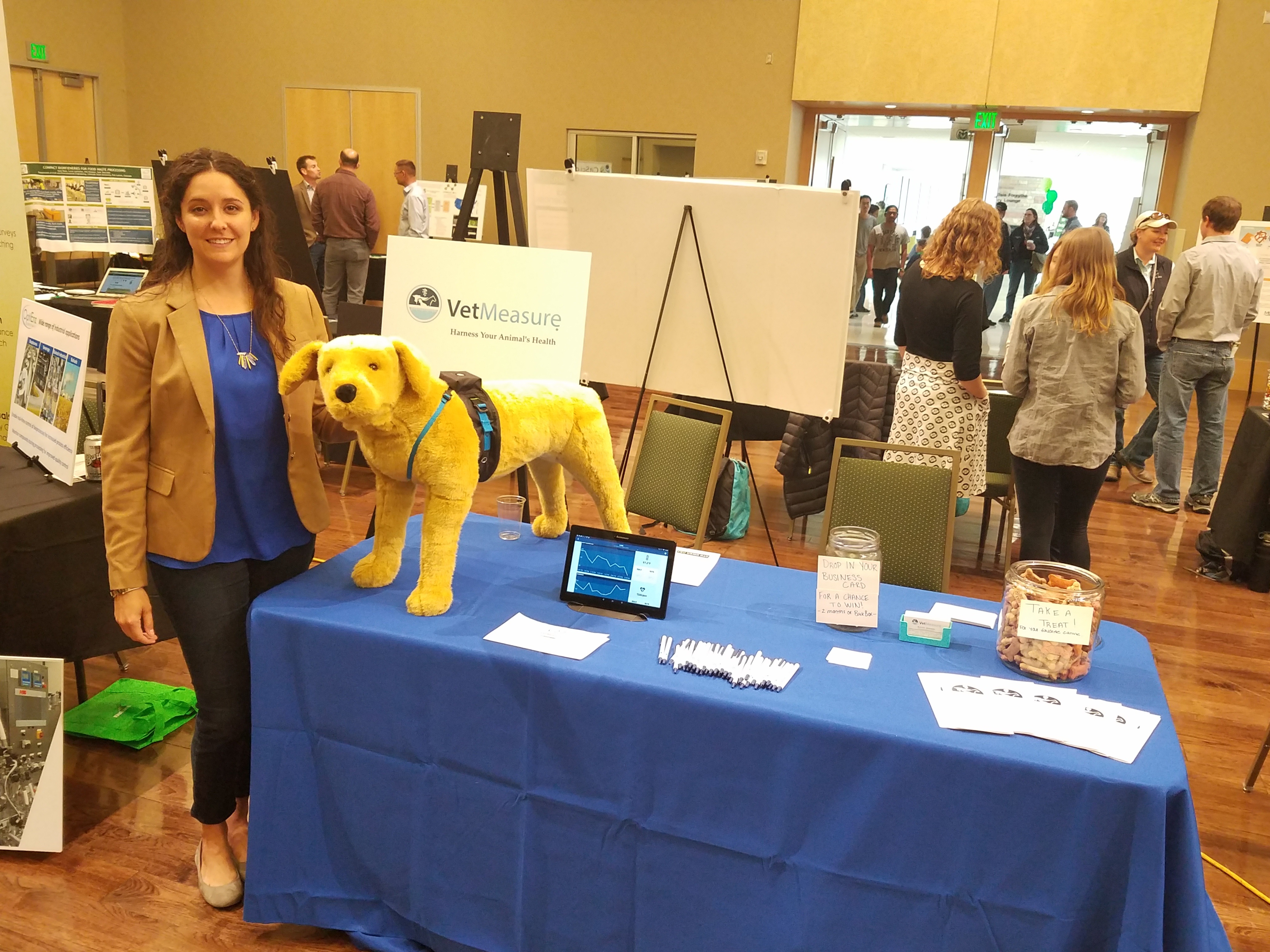 A female VetMeasure representative wearing black pants, a royal blue shirt, and a tan jacket stand next to a trade-show table of VetMeasure promotional items. Additional tables and people are visible in the background. 